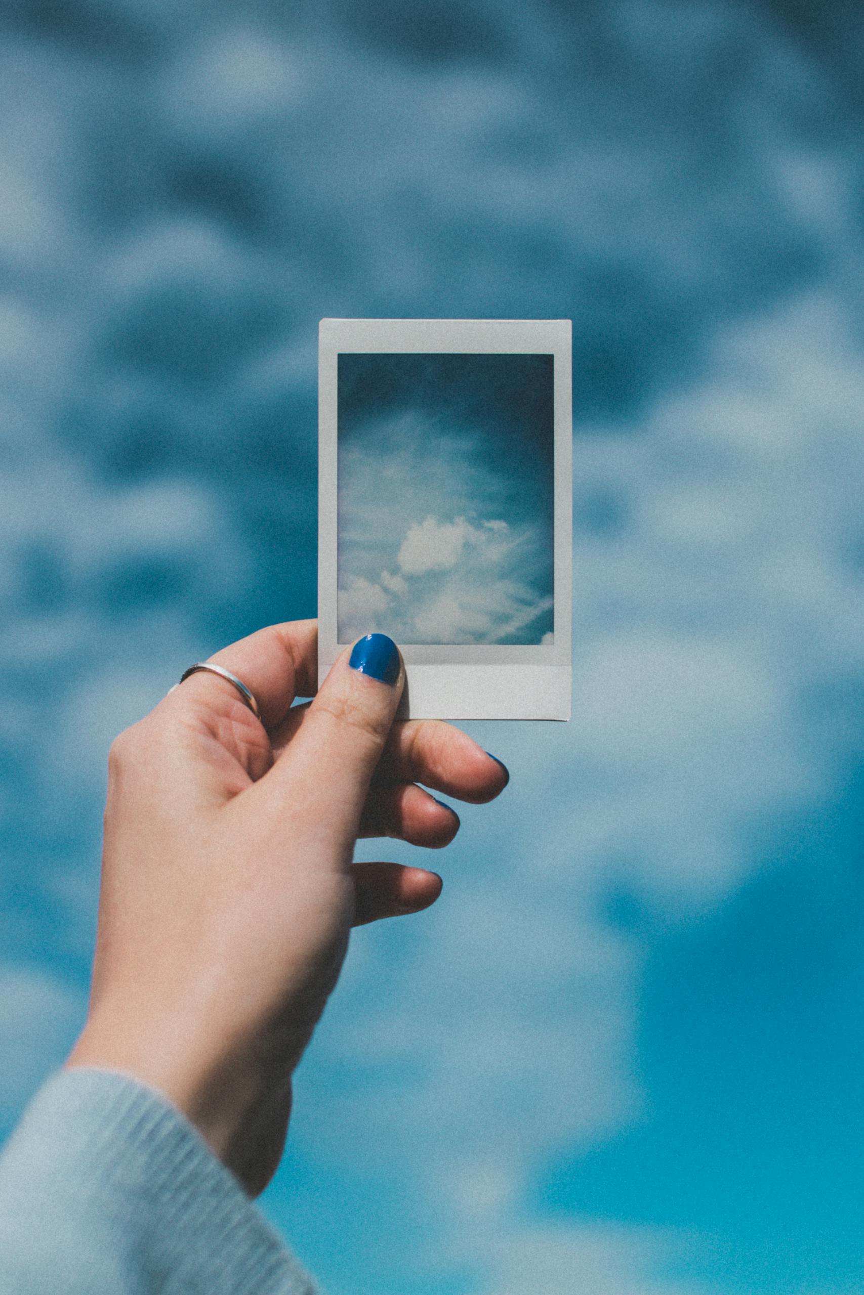A hand with painted nails holds a Polaroid picture of clouds against a vibrant blue sky background.